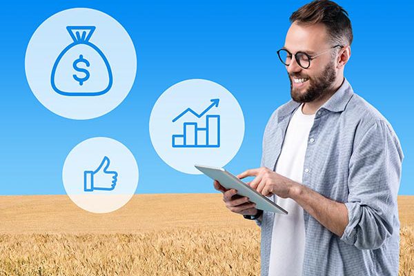 Millennial agribusinessman farmer using digital tablet for agribusiness processes automatization, icons with dollar bag, thumb up and financial bar, wheat field background, creative image